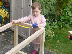Laylaaged two carefully measuring a handmade wooden toy chest frame with a tape measure in the garden — an early moment of hands-on learning and father-daughter craftsmanship